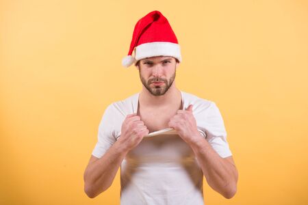 Macho with beard on serious face in red xmas hat. Man santa in white tshirt on orange background. Santa claus fashion. Christmas and new year party. Holidays celebration concept.の写真素材