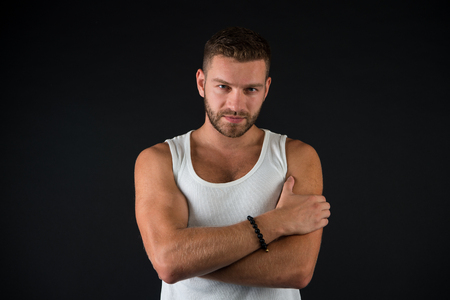 Man with bearded face and stylish hair on black background. Macho in white vest with folded muscular hands. Fitness, health, masculinity. Fashion, style, trend. Barbershop, male beauty conceptの写真素材