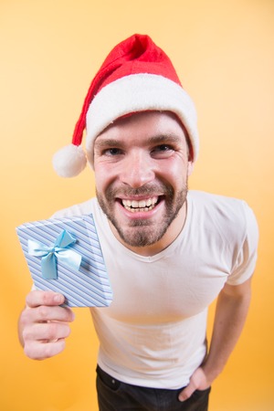 Gift giving, holiday greeting. Man in santa hat smile with present on orange background. Boxing day concept. Christmas, new year preparation and celebration. Macho hold xmas box with blue ribbon.の写真素材