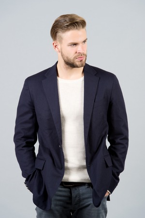 Macho in black jacket pose with hands in jeans pockets. Man with beard on unshaven face on grey...