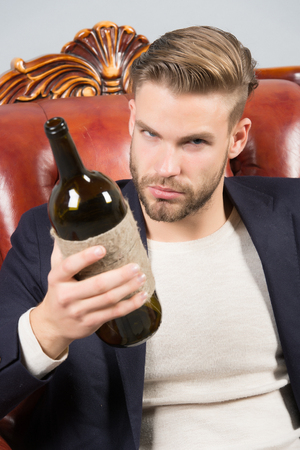 Man degustator with beard, stylish hair hold wine bottle on brown leather armchair. Wine tasting, degustation concept. Alcohol addiction, bad habits.の写真素材