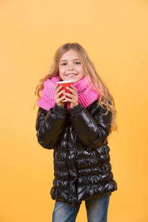 Autumn season concept. Girl with long blond hair in black jacket hold mug. Tea or coffee take away. Child smile with red cup on orange background. Warm up drink.の写真素材