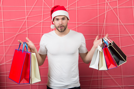 Man santa with shopping bags on pink background. Christmas, new year gift, present. Macho shopper in red hat hold paperbags. xmas sale concept. Winter holidays celebration.の写真素材