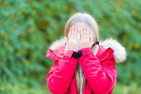 Girl hide face with hands on idyllic autumn day. Kid fashion and style. Happy childhood concept. Beauty, look, hairstyle. Child with blond hair ponytail in red coat on natural landscape.の写真素材