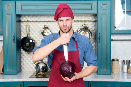 Man in red chef hat, apron cut purple cabbage with knife in kitchen. Cooking, culinary, cuisine. Vegetarian, diet, health. Food preparation conceptの写真素材