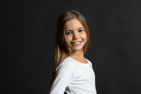 Happy child, childhood concept. Girl with smile on adorable face on dark background. Beauty, look, hairstyle. Kid model smiling with long healthy hair. Youth, skincare, health, copy spaceの写真素材