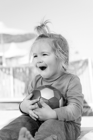 small baby boy or cute child with happy face and blonde hair in green shirt and pants sitting barefoot, playing with colorful ball at sunny summer outdoor beach on natural sky backgroundの写真素材