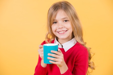Child smile with blue cup on orange background. Girl with long blond hair in red sweater hold mug. Thirst, dehydration concept. Health and healthy drink. Tea or coffee break.の写真素材