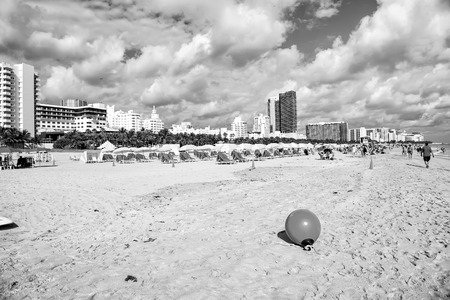 Miami, USA - January 10, 2016: south or miami beach. sand beach with deck chair under yellow umbrella, people, green palm and city skyline on sunny cloudy blue sky. Summer vacation. Lounge, sea resortのeditorial素材