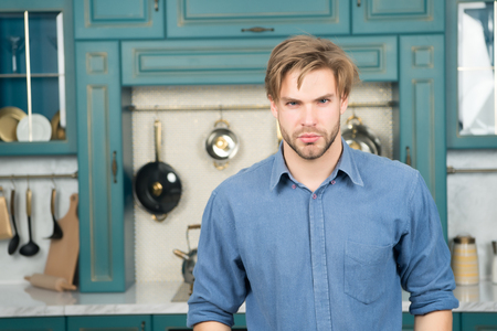 Man with serious face, blond hair in blue shirt stand in kitchen. Food preparation, cuisine, cooking concept.の写真素材