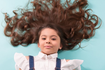 Girl with long healthy brunette hair on blue background. Little child with curly hairstyle, curls. Beauty or hairdressing salon. Haircare, health concept. Punchy pastel trendの写真素材