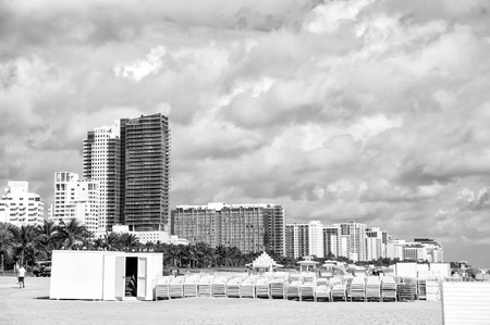 sunbeds stacked on sand beach, green palm trees and high rise, apartment buildings or hotels on sunny day. miami south beach skyline with white clouds on blue sky. Summer vacation. Urban landscapeのeditorial素材