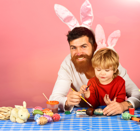 funny easter egg Easter celebration and joy concept. Father and son preparing for holiday. Man with beard and little boy painting eggs for Easter on pink background. Dad with painted nose wears pinkの写真素材