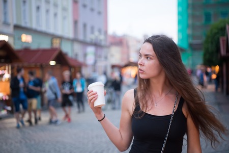 Girl with long hair walk on evening city street, travelling. Woman with disposable cup, coffee mood. Travelling, travel, trip. Coffee mood, recipe, takeaway drink. Vacation, adventure, discovery.の写真素材
