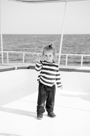 fashionable small baby boy or cute child with blonde hair in fashion stripped marine shirt and stylish pants on yacht white color outdoor on natural background with blue sea or ocean waterの写真素材