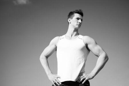 Man bodybuilder posing on blue sky. Athlete in green tshirt on sunny day. Healthy lifestyle concept. Fitness model with muscular hands, biceps, triceps, muscles. Workout and training, copy spaceの写真素材