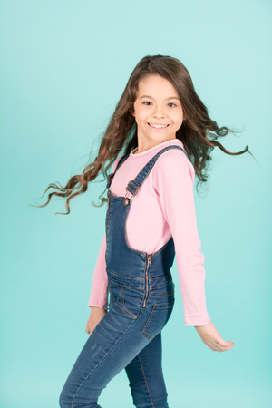 Child happy smiling with flowing long brunette hair, beauty. Little girl pose in jeans overall, fashion. Beauty, look, hairstyle. Fashion, casual style. Childhood, youth, punchy pastel trendの写真素材