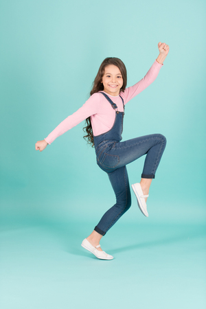Happy child in jeans overall dance, fashion. Small girl with long brunette hair dancing, activity. Fashion, casual style. Beauty, look, hairstyle. Activity, energy, fun, punchy pastel trendの写真素材