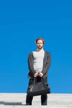 Bearded man with bag on blue sky, future. Macho with travelling bag on sunny outdoor, vacation. Future, freedom, success. Guy model ready for vacation, travel. Mens fashion with accessory, styleの写真素材