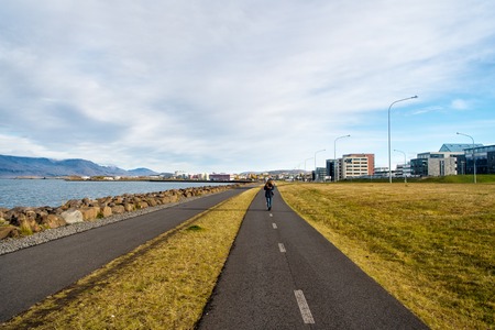 Woman walk on road along sea on cloudy sky in reykjavik, iceland. Promenade at seaside on cityscape. Travel and wanderlust on urban landscape. Freedom, perspective and future.の写真素材