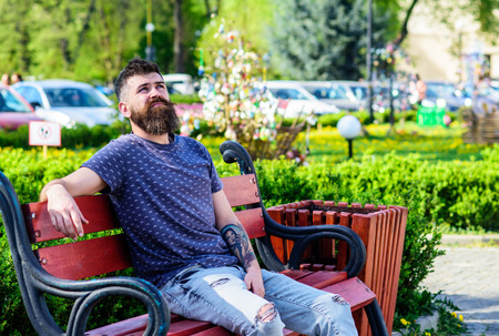 Man with beard and mustache on strict face sits on bench in park. Bearded man with fresh haircut relaxing, urban background. Public recreation places concept. Hipster enjoy sunny day in parkの写真素材