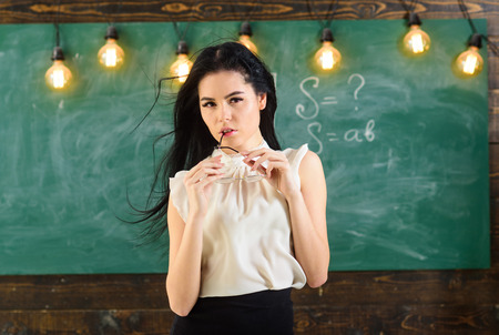 Woman with long hair in white blouse stands in classroom. Teacher with glasses and waving hair looks sexy. Sexy teacher concept. Lady strict teacher on dreamy face stands in front of chalkboardの写真素材