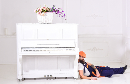 Loader lean on piano instrument. Lazy worker concept. Man with beard, worker in overalls and helmet fall asleep tired , white background. Courier fall asleep while moveing furniture, relocation.の写真素材