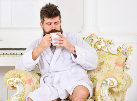 Man sleepy in bathrobe, drinks coffee, enjoying aroma in luxury hotel in morning, white background. Man with beard and mustache enjoys morning while sitting on luxury armchair. Elite leisure conceptの写真素材