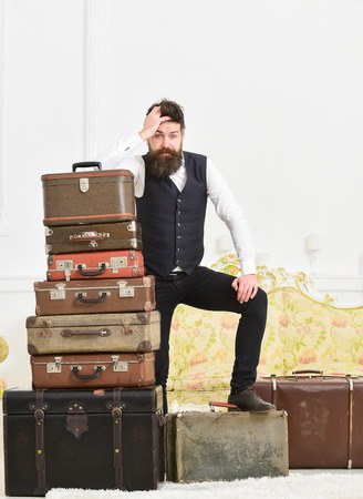 Macho elegant on disappointed face standing near pile of vintage suitcase. Man, butler with beard and mustache delivers luggage, luxury white interior background. Luggage and travelling conceptの写真素材
