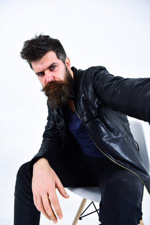 Man with beard and mustache on strict face looks at camera. Macho wears leather jacket, white background. Menswear and fashion concept. Hipster looks serious while sitting on chair in stylish outfitの写真素材