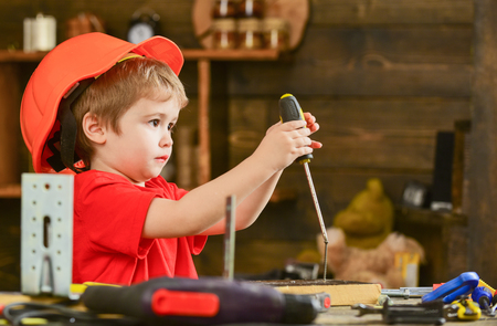 Side view little kid binding screw into wooden block. Concentrated boy in orange helmet sitting at table and holding screwdriverの写真素材