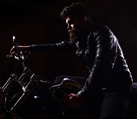 Macho, brutal biker in leather jacket riding motorcycle at night time, copy space. Man with beard, biker in leather jacket sitting on motor bike in darkness, black background. Night racer conceptの写真素材