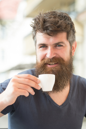 Bearded man holds espresso cup, drinks coffee. Coffee break concept. Man with long beard looks cheerful and relaxed. Man with beard and mustache on happy smiling face, light background, defocusedの写真素材