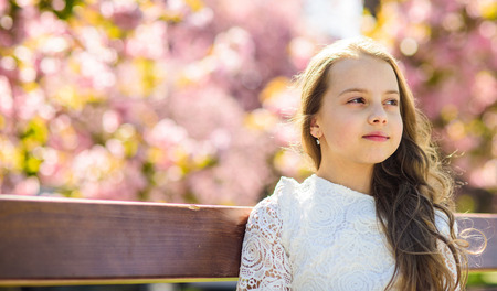 Girl on dreamy face sits on bench, sakura tree on background, defocused. Cute child with long beautiful hair enjoy sunny spring day. Girl relaxing in park, copy space. Natural beauty conceptの写真素材