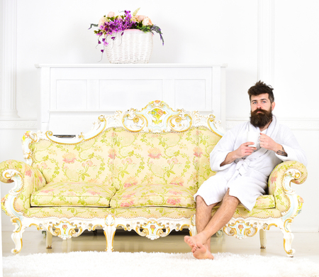 Elite leisure concept. Man on sleepy face in bathrobe, drinks coffee, in luxury hotel in morning, white background. Man with beard and mustache enjoys morning while sitting on luxury sofaの写真素材