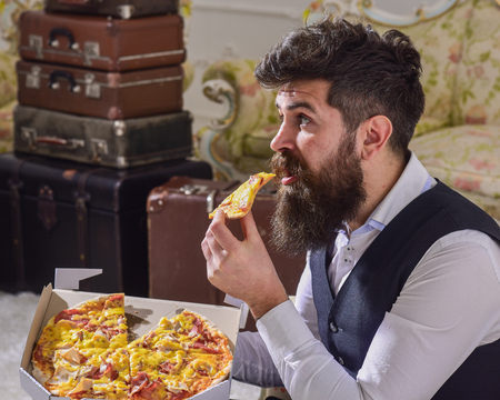 Man with beard and mustache holds delivered box with tasty fresh hot pizza. Pizza delivery concept. Macho in classic clothes hungry, on surprised face, bites slice of pizza, eats, interior backgroundの写真素材
