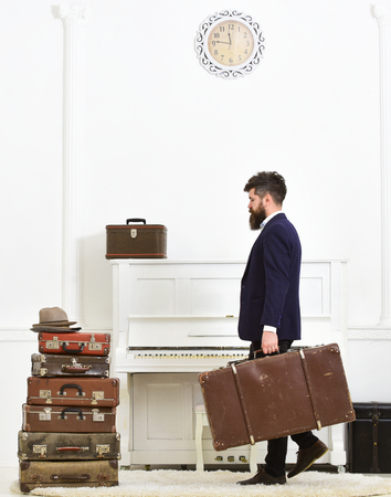 Butler and service concept. Macho attractive, elegant carries vintage suitcases, side view. Man with beard and mustache wearing classic suit delivers luggage, luxury white interior backgroundの写真素材