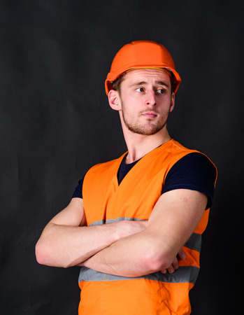 Man in helmet, hard hat hold arms crossed on chest, black background. Worker, contractor, builder on strict face with muscular biceps. Builder in helmet posing confidently. Strong builder conceptの写真素材