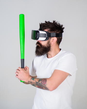 Man with beard in VR glasses, grey background. VR sportsman concept. Hipster on concentrated face use modern technologies for sporty entertainment. Guy with VR glasses play baseball with baseball batの写真素材