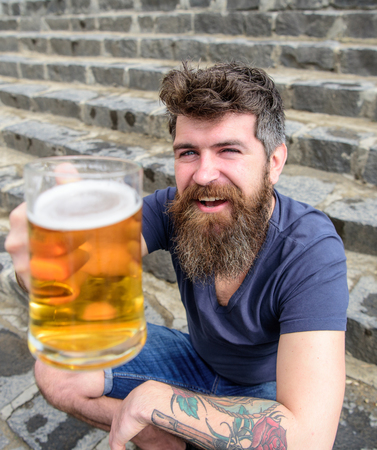 Hipster on cheerful face drinks beer outdoor. Celebration concept. Man with beard and mustache holds...