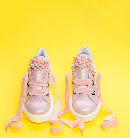 Footwear for girls or women decorated with pearl beads. Pair of pale pink female sneakers with velvet ribbons. Cute shoes on yellow background. Childrens shoes conceptの写真素材