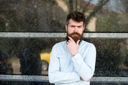 Brutal man with serious face touching his beard while thinking about something. White-collar man standing outside his office building, break at workの写真素材