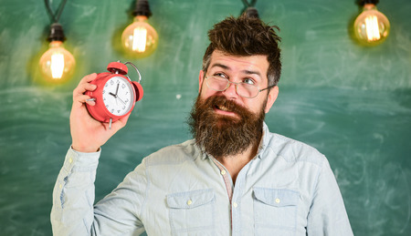 Teacher in eyeglasses holds alarm clock. Man with beard and mustache on annoyed face listens alarm...