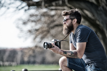 Bearded man works with vintage camera. Man with beard and mustache on concentrated face, branches on background, defocused. Man with long beard busy with shooting photos. Photographer conceptの写真素材