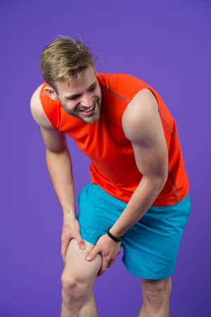 Sportsman with knee pain on violet background. Man in orange vest and blue shorts. Health problem...