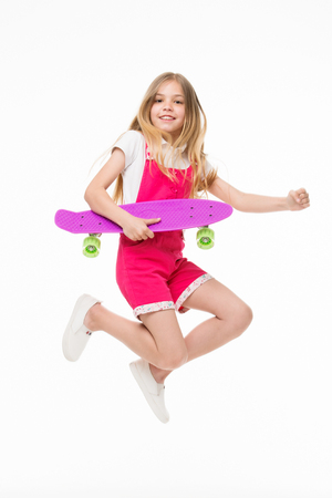 Find your freedom! Small girl jump with skate board isolated on white. Child skater smile with longboard. Skateboard kid in pink jumpsuit. Sport activity and energy. Childhood and active games.の写真素材