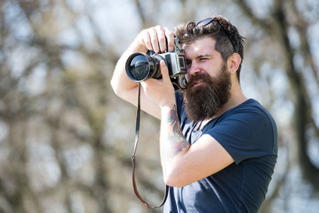 Young male photographer taking pictures in the park with vintage camera, hobby and leisure activity concept. Bearded hipster searching for best spot for photo shootingの写真素材