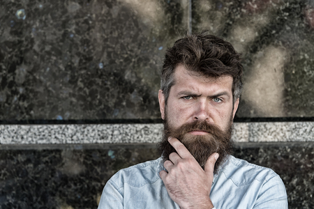 Man with beard and mustache on concentrated face, black marble background. Masculinity concept. Hipster with tousled hair touches beard while looking at camera. Guy looks interested and curiousの写真素材