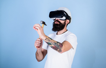 Builder and renovation concept. Man with beard in VR glasses holds hammer, light blue background. Guy with HMD hammering virtual nail into VR surface. Hipster busy with renovation in virtual realityの写真素材