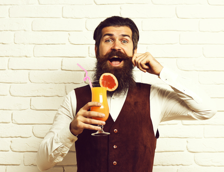 handsome bearded man with long beard and mustache has stylish hair on happy face holding glass of alcoholic beverage in vintage suede leather waistcoat on white brick wall studioの写真素材